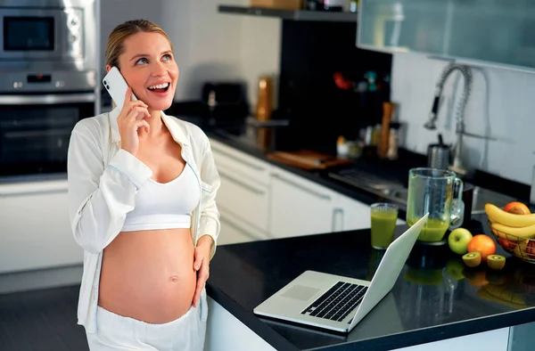 一个年轻的孕妇在厨房里的笔记本电脑前工作 在电话里聊天 在一张刚刚准备好的维生素冰沙的桌子上 怀孕期间的工作和健康饮食 — 图库照片