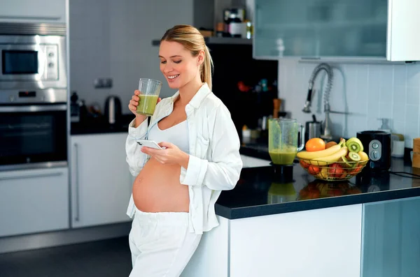 一个年轻的孕妇喝着准备好的维生素冰沙 在厨房里用智能手机站在家里 怀孕期间的健康饮食 — 图库照片