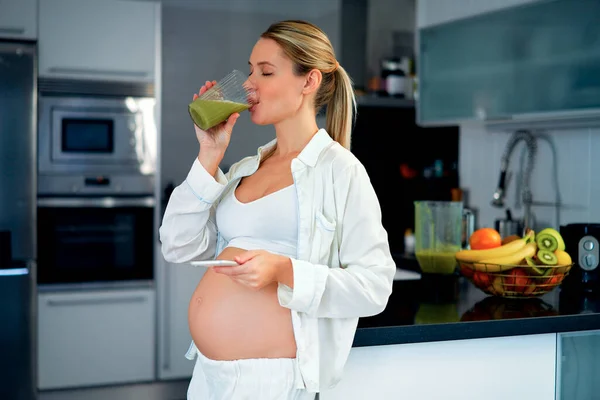 一个年轻的孕妇喝着准备好的维生素冰沙 在厨房里用智能手机站在家里 怀孕期间的健康饮食 — 图库照片