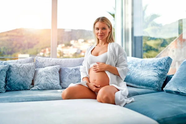 一个穿着白色外套 双手放在肚子上的年轻孕妇坐在她家窗边一个天鹅绒的蓝色沙发上 母亲和怀孕的概念 — 图库照片