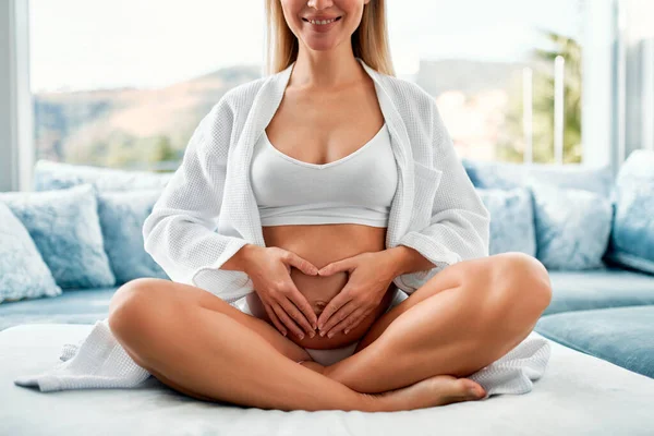 一个穿着白色外套 双手放在肚子上的年轻孕妇坐在她家窗边一个天鹅绒的蓝色沙发上 母亲和怀孕的概念 — 图库照片