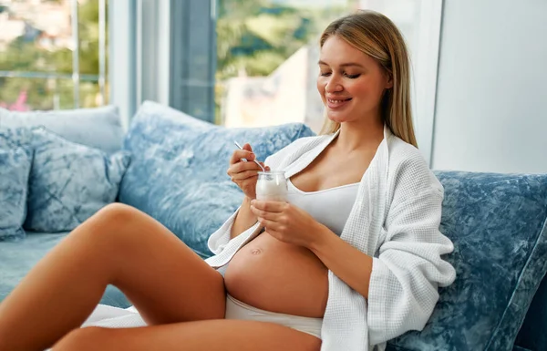 迷人的孕妇正坐在靠窗的蓝色天鹅绒沙发上 吃着酸奶 健康食品概念 怀孕的最后几个月 — 图库照片