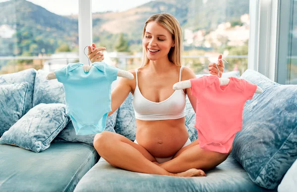 一个穿着粉色和蓝色婴儿衣服的漂亮的年轻孕妇坐在家里的沙发上 猜谁会生男孩或女孩 母亲和怀孕的概念 — 图库照片