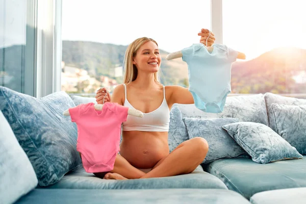一个穿着粉色和蓝色婴儿衣服的漂亮的年轻孕妇坐在家里的沙发上 猜谁会生男孩或女孩 母亲和怀孕的概念 — 图库照片