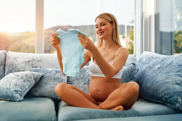 一位年轻的孕妇抱着蓝色的婴儿衣服 坐在客厅里她家窗边的蓝色天鹅绒沙发上 怀孕和生育的概念 — 图库照片