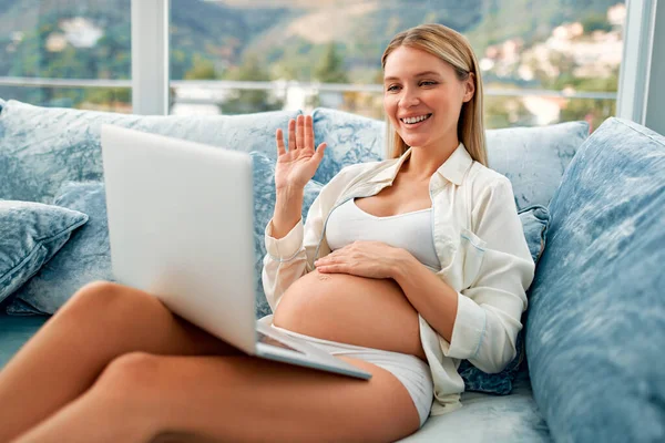一位年轻的孕妇坐在沙发上 手里拿着笔记本电脑 挥动着手 在客厅里的家中进行视频通话 产妇和怀孕概念 — 图库照片