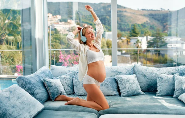 A young beautiful pregnant woman in white underwear and a bathrobe with headphones listening to music and dancing kneeling on a blue velvet sofa in the living room at home. The concept of pregnancy and motherhood.
