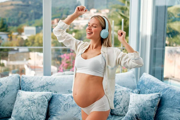 A young beautiful pregnant woman in white underwear and a bathrobe with headphones listening to music and dancing kneeling on a blue velvet sofa in the living room at home. The concept of pregnancy and motherhood.