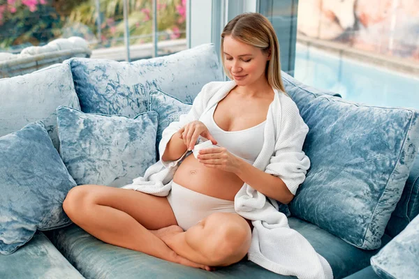 一位穿着白色内裤和浴衣的年轻孕妇坐在家中客厅的沙发上 用保湿霜擦拭肚子的皮肤 母亲和怀孕的概念 — 图库照片