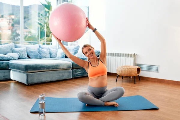 一位穿着运动服的年轻孕妇坐在客厅的橡胶垫子上 带着一个健身球做运动 怀孕期间的运动和健康 — 图库照片
