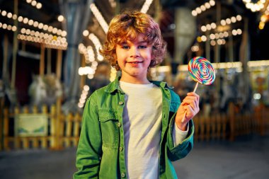 Sarışın, kıvırcık saçlı, atlıkarıncaların arka planında akşam vakti bir lunaparkta ya da sirkte duran renkli bir lolipop yiyen beyaz bir çocuk..