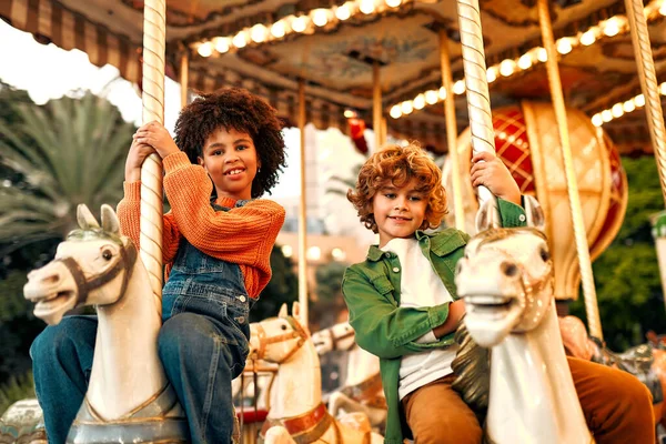An African-American girl with Afro-curls and a Caucasian curly-haired blonde boy ride a vintage horse carousel in the evening at an amusement park on a weekend.