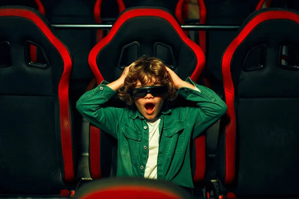 A cute Caucasian boy with curly hair sitting in a virtual cinema in glasses on moving rotating chairs and emotionally watching a movie in an amusement park.