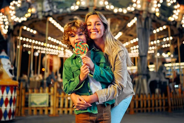 Annesi ile birlikte sarı kıvırcık saçlı, beyaz bir çocuk atlıkarıncanın arka planında duran renkli lolipopu yiyor. Akşamları lunaparkta ya da sirkte..