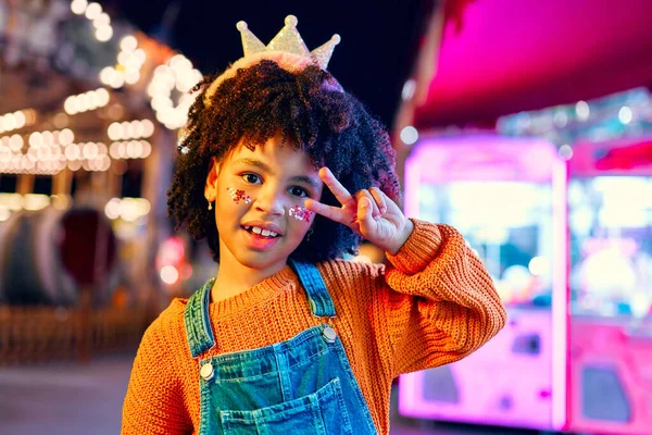 A cute African-American girl with an Afro hairstyle, sequins on her cheeks and a crown on her head like a princess standing in front of glowing carousels at an amusement park in the evening.