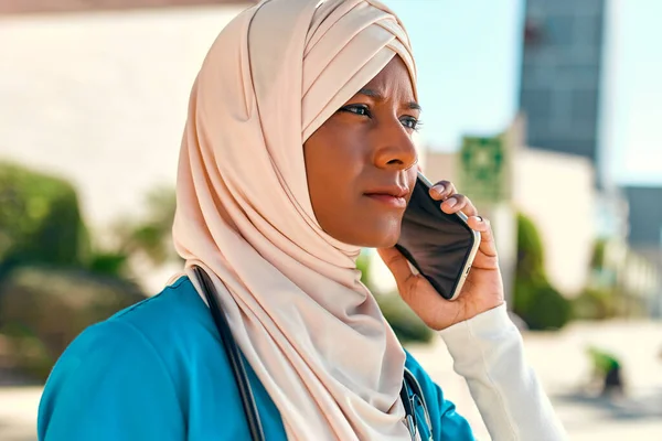 Young Muslim woman in hijab doctor in uniform with a stethoscope talking on the phone while standing on the street near the hospital building. Medicine and health care.