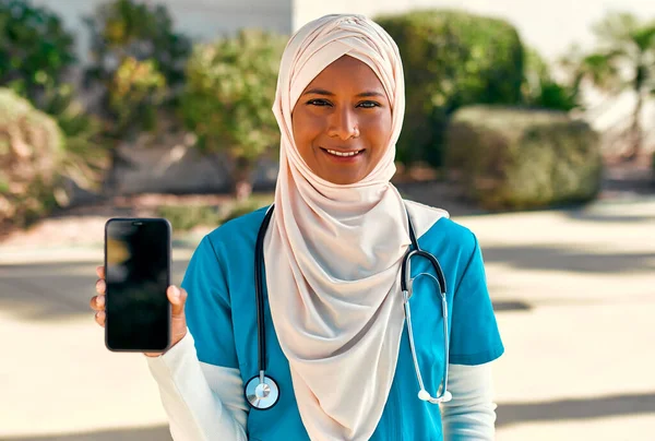 Young Muslim woman in hijab doctor in uniform with stethoscope showing smartphone screen while standing on the street near the hospital building. Medicine and health care.