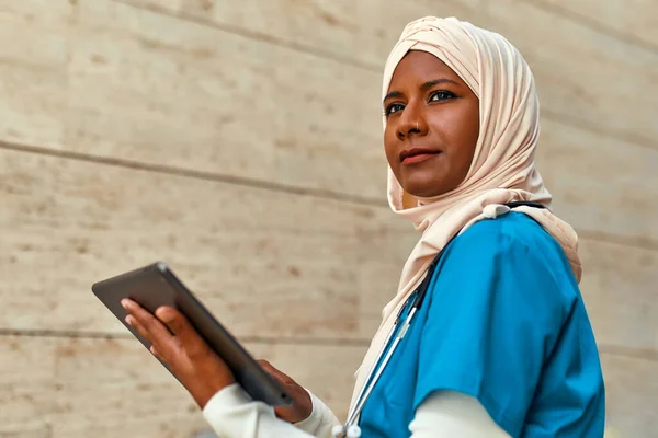 Young Muslim woman in hijab doctor in uniform with a stethoscope holding a tablet with x-rays standing on the street near the hospital building. Medicine and health care.
