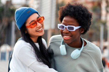 Afro-Amerikalı ve Asyalı genç tatlı bayan kız arkadaşlar kucaklaşıyorlar ve dışarıda kulaklıkla müzik dinliyorlar..