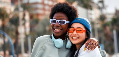 Afro-Amerikalı ve Asyalı genç tatlı bayan kız arkadaşlar kucaklaşıyorlar ve dışarıda kulaklıkla müzik dinliyorlar..