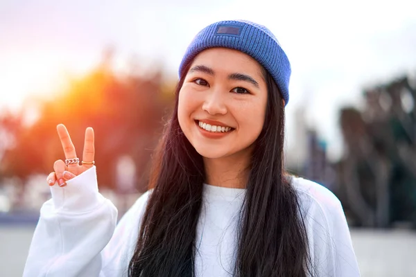 閉じるかわいいですアジアの若いです女性でキャップ立って屋外と笑顔ショー二本の指自由ジェスチャー — ストック写真