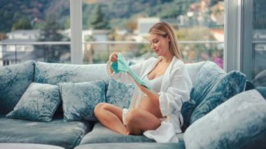 Evinin penceresinin yanındaki mavi kadife koltukta oturan mavi bebek kıyafetleri tutan hamile bir kadın. Hamilelik ve annelik kavramı..
