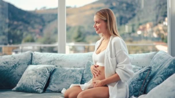 一个穿着白色外套 双手放在肚子上的年轻孕妇坐在她家窗边一个天鹅绒的蓝色沙发上 母亲和怀孕的概念 — 图库视频影像