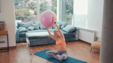 Spor kıyafetli genç hamile bir kadın evin oturma odasındaki lastik paspasın üzerinde spor topuyla egzersiz yapıyor. Hamilelik sırasındaki spor.