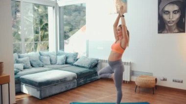 Spor giyimli hamile bir kadın evin oturma odasındaki lastik bir paspasın üzerinde egzersiz yapıyor. Hamilelik boyunca spor ve yoga.