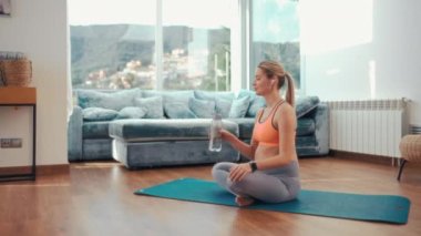 Spor kıyafetli genç hamile bir kadın lastik bir paspasın üzerinde oturuyor ve evin oturma odasındaki egzersizden sonra su içiyor. Hamilelik sırasındaki spor.