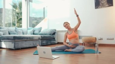 Spor kıyafetli genç hamile bir kadın evdeki oturma odasında lastik bir paspasın üzerinde oturmuş dizüstü bilgisayardaki video derslerinde yoga sporları yapıyor. Hamilelik süresince aktif yaşam tarzı.