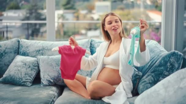 一个穿着粉色和蓝色婴儿衣服的漂亮的年轻孕妇坐在家里的沙发上 猜谁会生男孩或女孩 母亲和怀孕的概念 — 图库视频影像