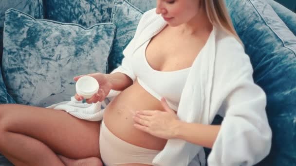 一位穿着白色内裤和浴衣的年轻孕妇坐在家中客厅的沙发上 用保湿霜擦拭肚子的皮肤 — 图库视频影像