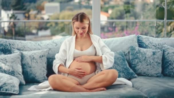 白いコートに身を包んだ若い妊婦が 家の窓辺の青いソファに腰を下ろしている 母親と妊娠の概念 — ストック動画