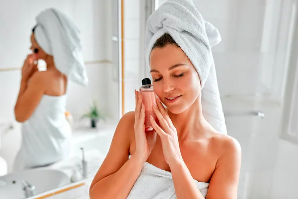 一位金发女郎穿着毛巾站在镜子前的浴室里 每天早上做面部和身体皮肤护理 手里拿着一瓶胶水或乳液 化妆品和温泉疗程 — 图库照片