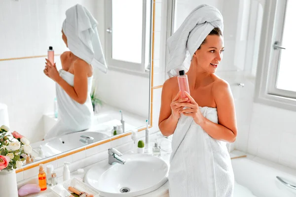 鏡の前の浴室に立つタオルのブロンドの女性は 顔とボディスキンケアのための彼女の朝のルーチンを実行し ゲルやローションのボトルを保持します 美容とスパの手順 — ストック写真