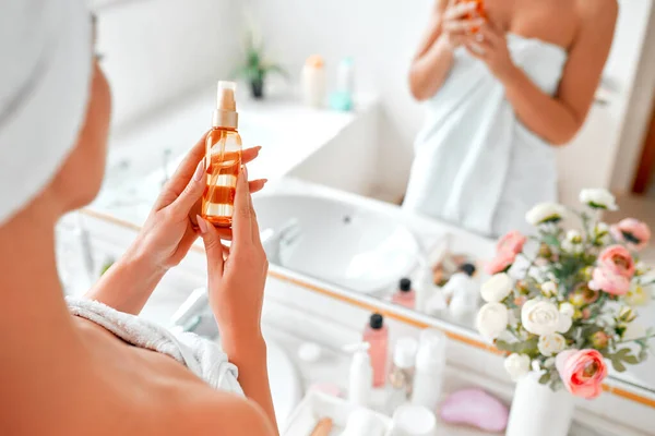 A blonde woman in a towel standing in the bathroom at the mirror, doing her morning routine for face and body skin care using oil or serum. Cosmetology and spa procedures.