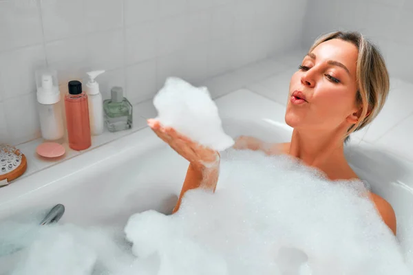 美しい女性は泡風呂に横たわって 彼女の手のひらから泡が吹いている 美容とスパの手順 — ストック写真