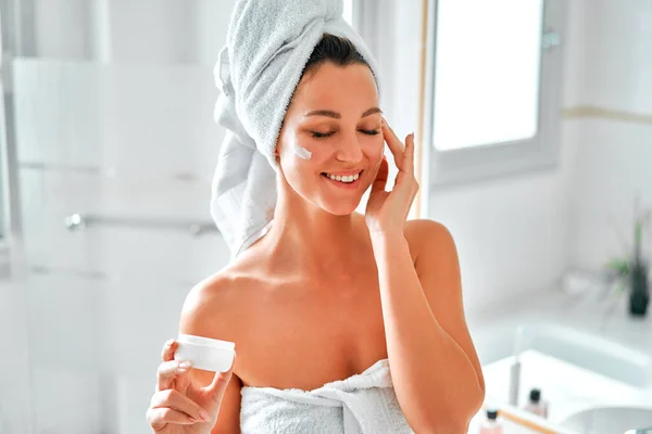 洗面台に立つ白いタオルに身を包んだ美しい女性と 保湿クリームを使った鏡 朝のスキンケアルーチン 美容とスパの手順 — ストック写真