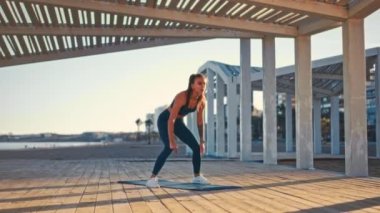 Young pretty fit woman in sportswear doing morning exercises on a rubber mat on the beach. Outdoor workout by the sea. Sports and recreation, fitness and healthy lifestyle.