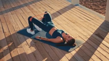 Young pretty fit woman in sportswear doing morning exercises on a rubber mat on the beach. Outdoor workout by the sea. Sports and recreation, fitness and healthy lifestyle.