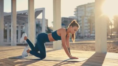 Young pretty fit woman in sportswear doing morning exercises on a rubber mat on the beach. Outdoor workout by the sea. Sports and recreation, fitness and healthy lifestyle.
