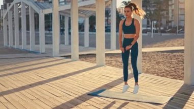 Young pretty fit woman in sportswear doing morning exercises on a rubber mat on the beach. Outdoor workout by the sea. Sports and recreation, fitness and healthy lifestyle.