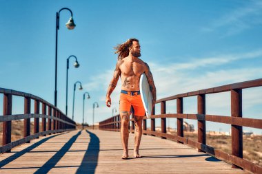 Yüzme şortlu, elinde sörf tahtası olan beyaz sakallı bir adam denize giden ahşap bir köprü boyunca yürüyor. Spor ve aktif eğlence..