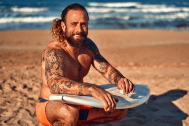 Yüzme şortlu, sakallı, beyaz bir adam deniz kenarında sörf tahtasını cilalıyor. Spor ve aktif eğlence..