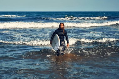 Kafkas kaslı, sakallı, uzun saçlı, dalgıç kıyafetli ve deniz kenarında sörf tahtası olan bir adam. Spor ve aktif eğlence..