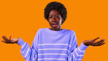 Afro saçlı, parlak turuncu arka planda mavi bir süveter giyen Afro-Amerikalı genç bir kadın inançsızlıkla kollarını açtı..