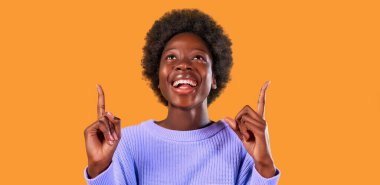 Afro saçlı, parlak turuncu arka planda mavi bir süveter giyen Afro-Amerikalı genç bir kadın reklam ürününü eklemek için başparmağını kaldırıyor..