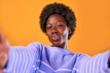 Afro saçlı, parlak turuncu arka planda mavi bir süveter giyen Afro-Amerikalı genç bir kadın kameraya selfie çekiyor..