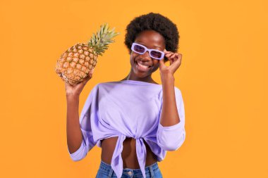 Afro saçlı, mor güneş gözlüğü takan, turuncu arka planda ananas tutan Afro-Amerikalı genç bir kadın..